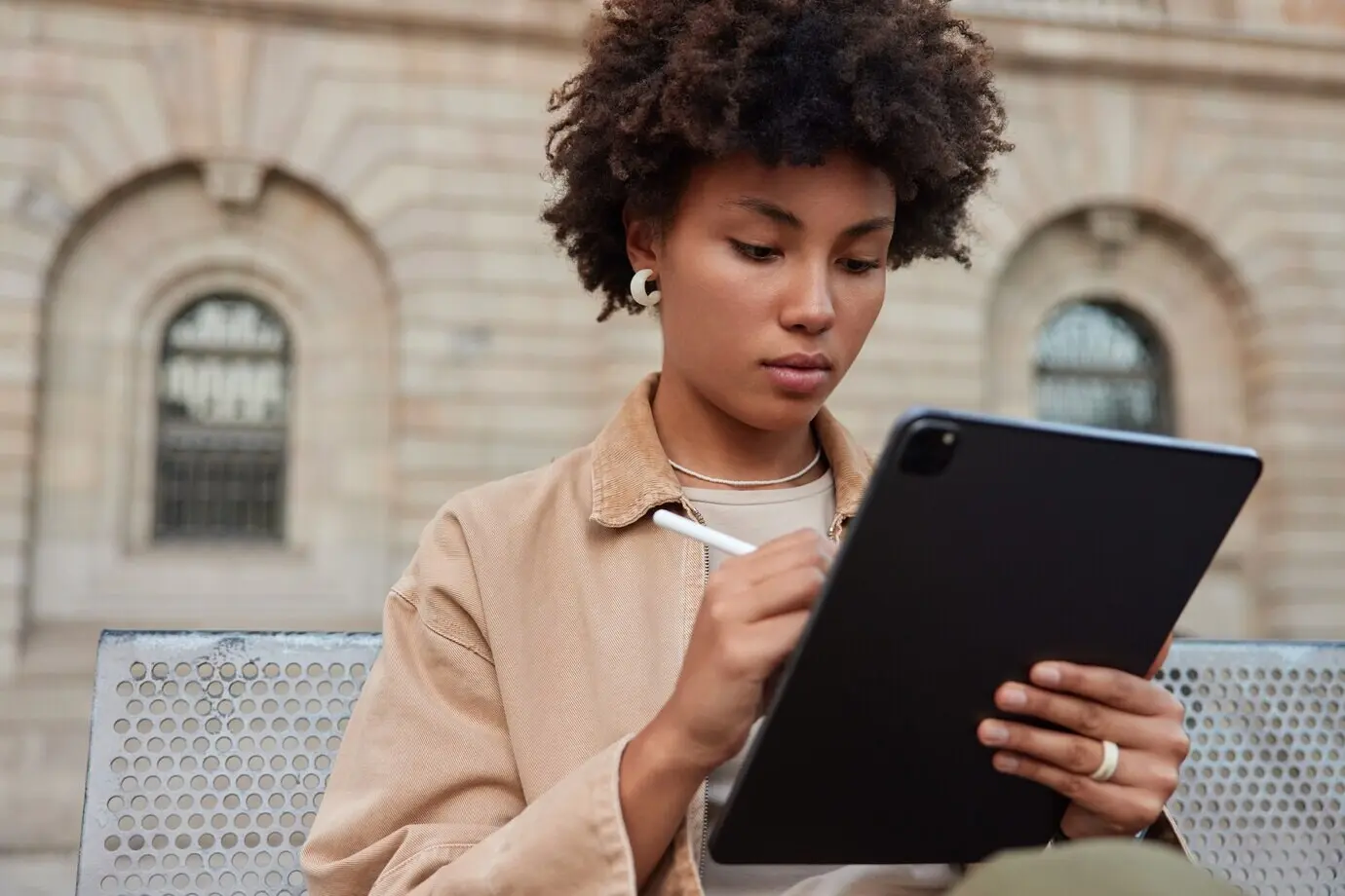 Im Freien sitzt eine stilvoll gekleidete Frau und arbeitet mit einem Eingabestift an einem Tablet: Sie erstellt Skizzen für ein bevorstehendes Projekt und bearbeitet Bilder für die Bildproduktion.
